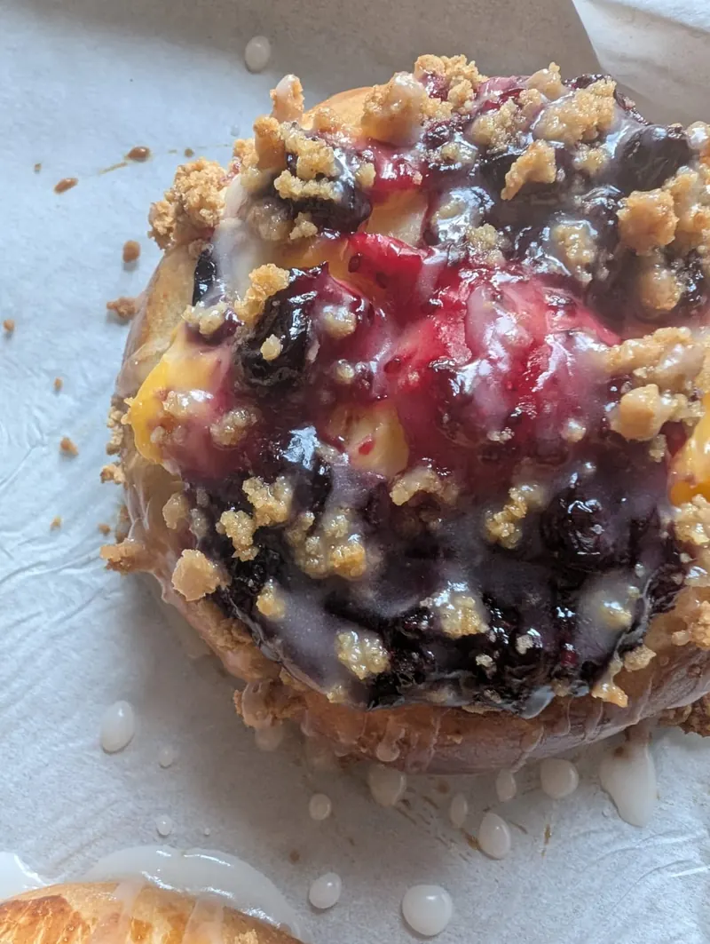 Close-up of a bun topped with glossy blueberries, raspberries, crumble and icing